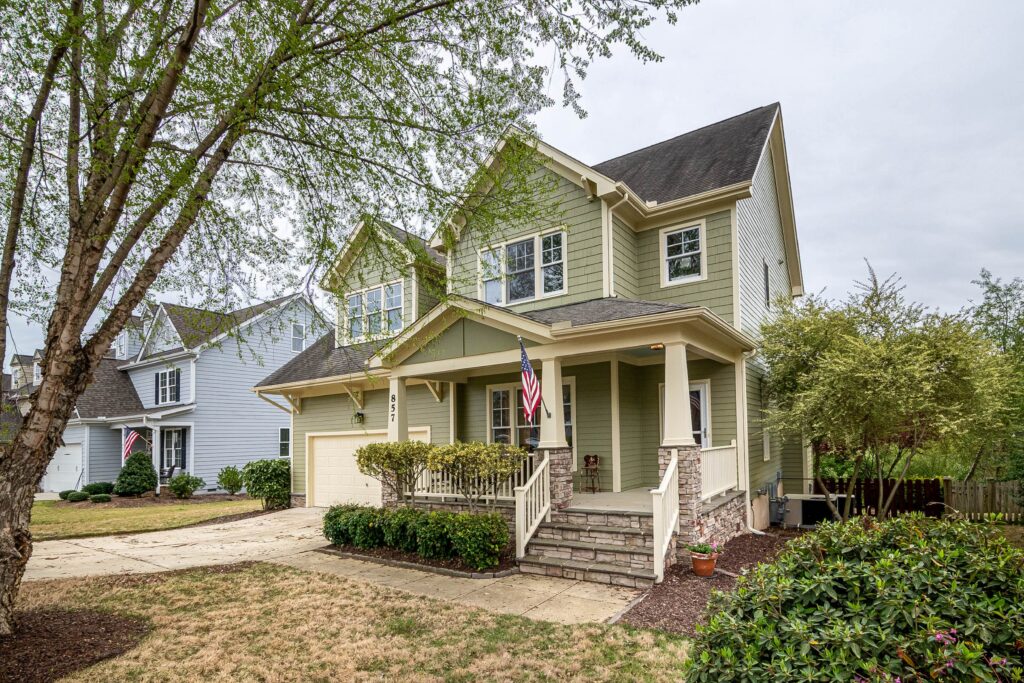 A green two story home with a porch, shingle roof and leafy yard. Illustrative stock property.